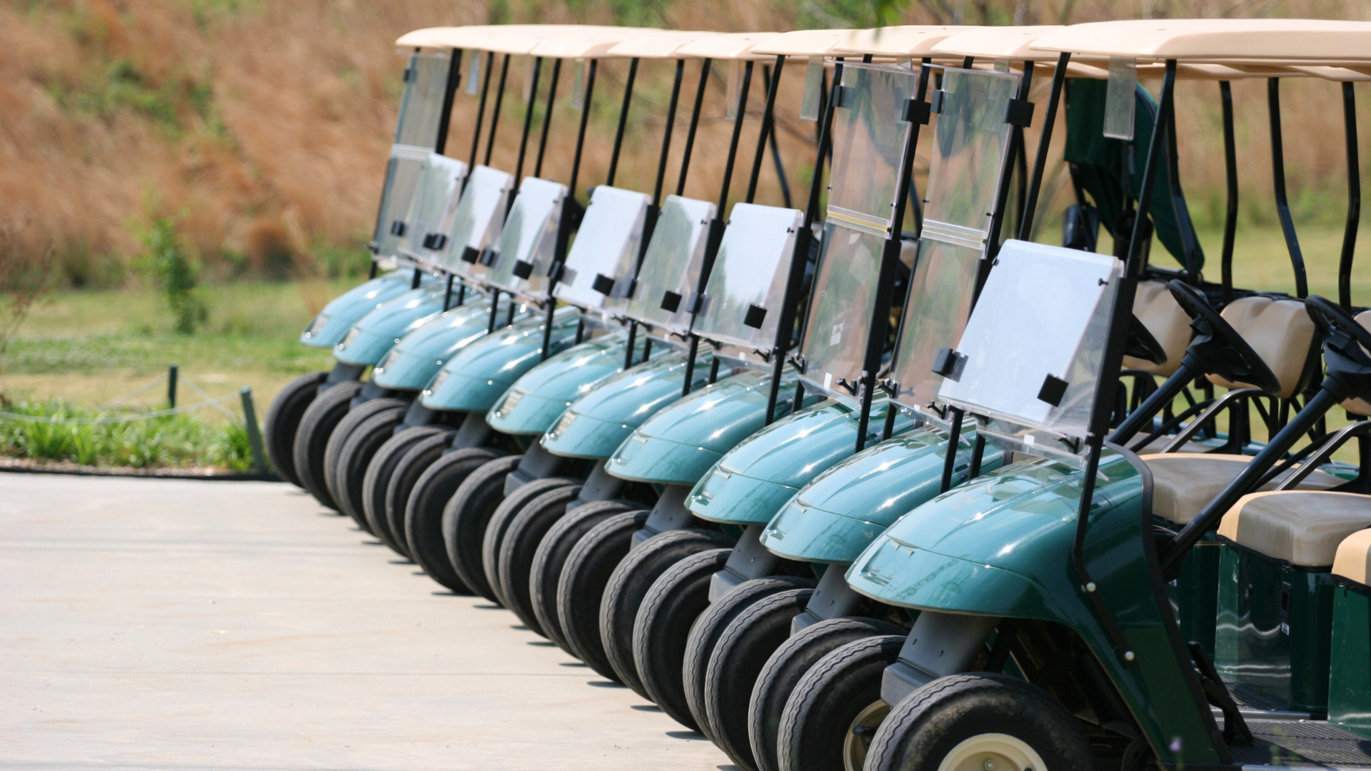 Row of green golf carts parked side by side outdoors with clear windshields and beige roofs.
