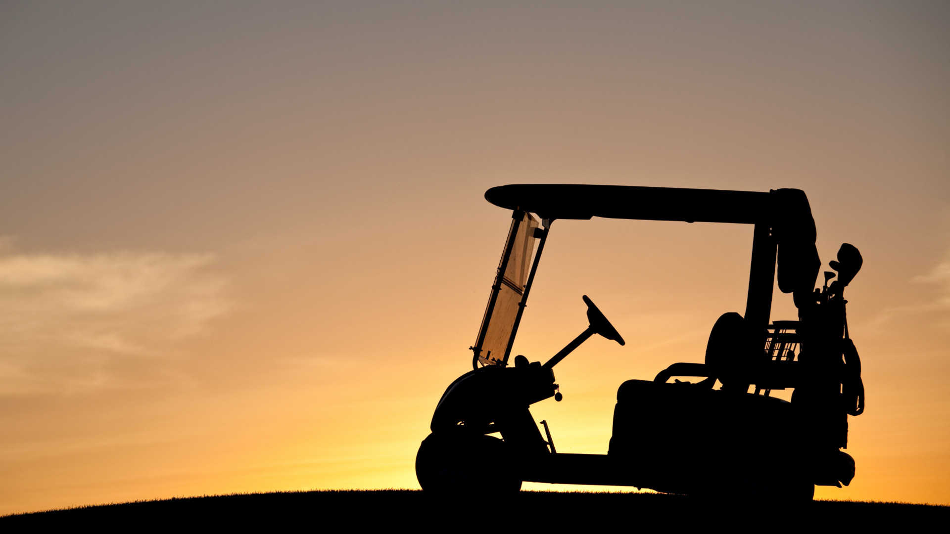 Silhouette of a golf cart on a hill at sunset with golf clubs mounted in back.