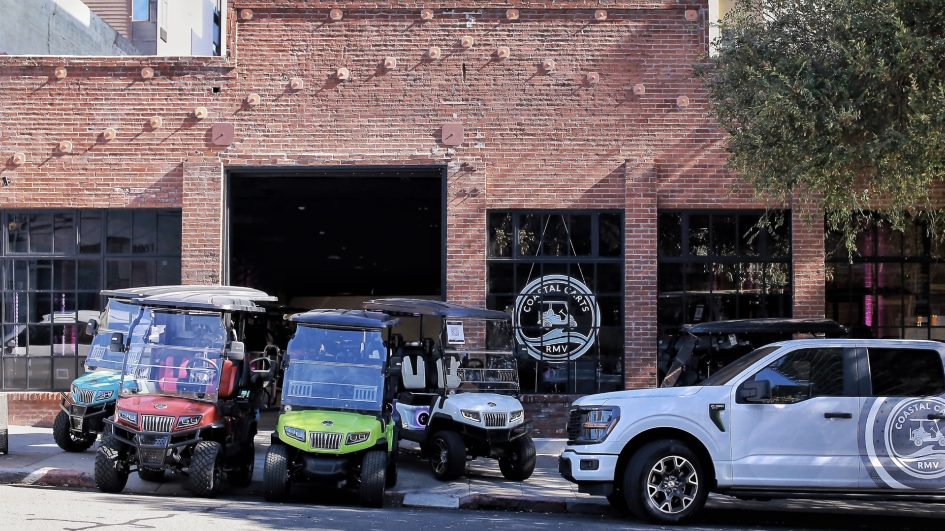 Custom golf carts and branded Coastal Carts RMV truck parked outside a brick storefront.