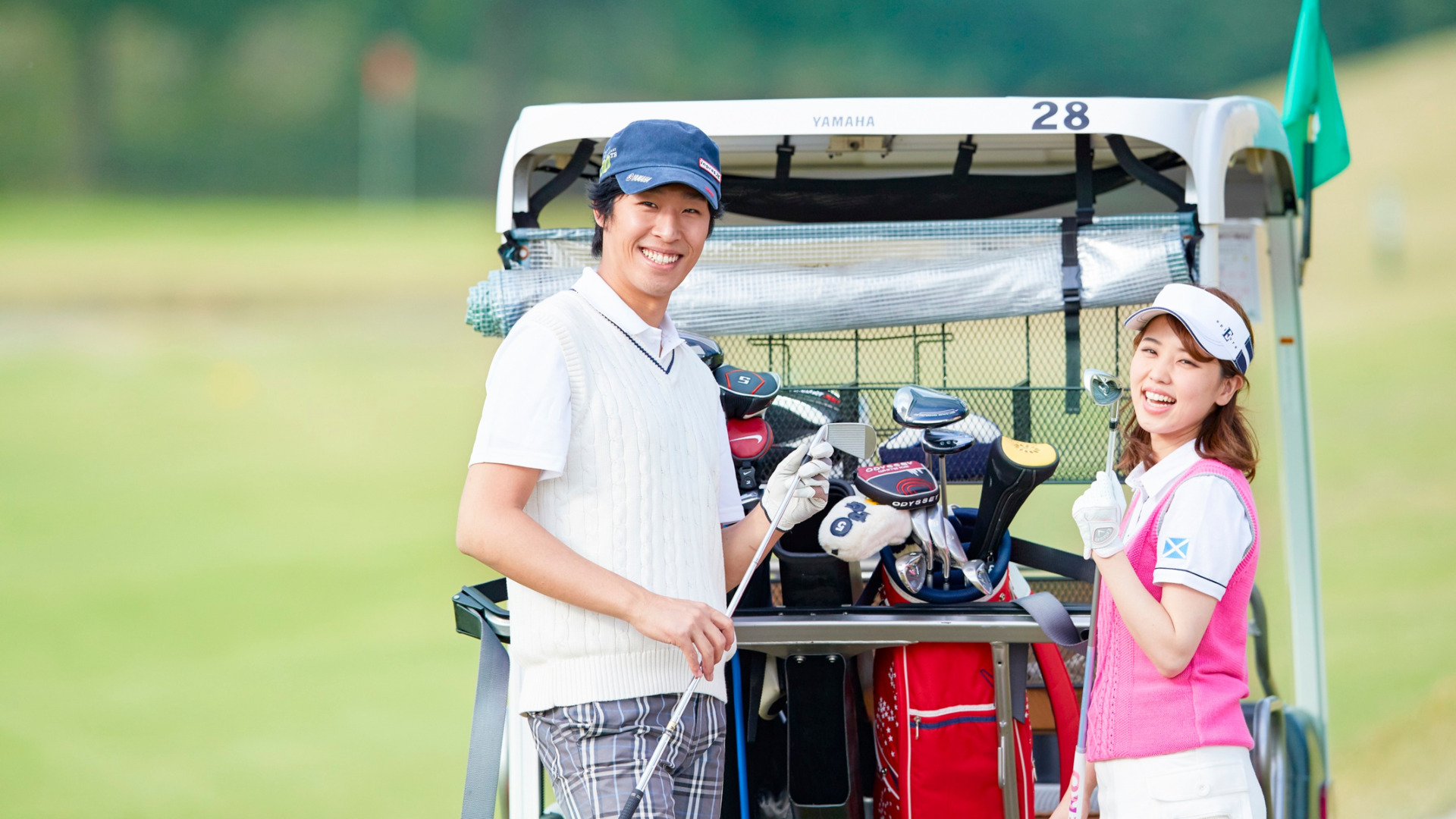Two golfers smiling beside a golf cart loaded with clubs on a golf course.