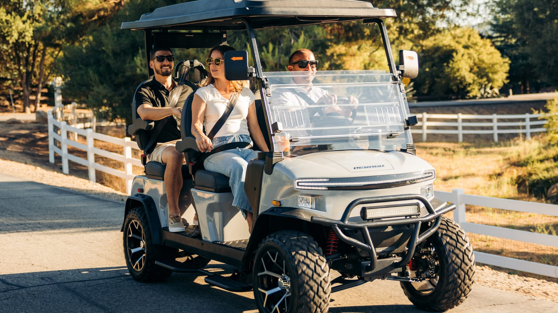 2026 Denago Rover XL white golf cart carrying four passengers on a paved road near a ranch fence.