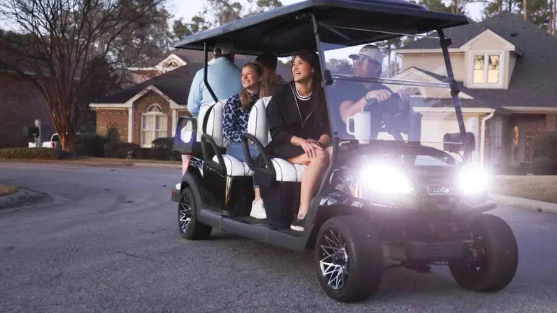 Black four-passenger Club Car golf cart driving through a neighborhood at dusk.