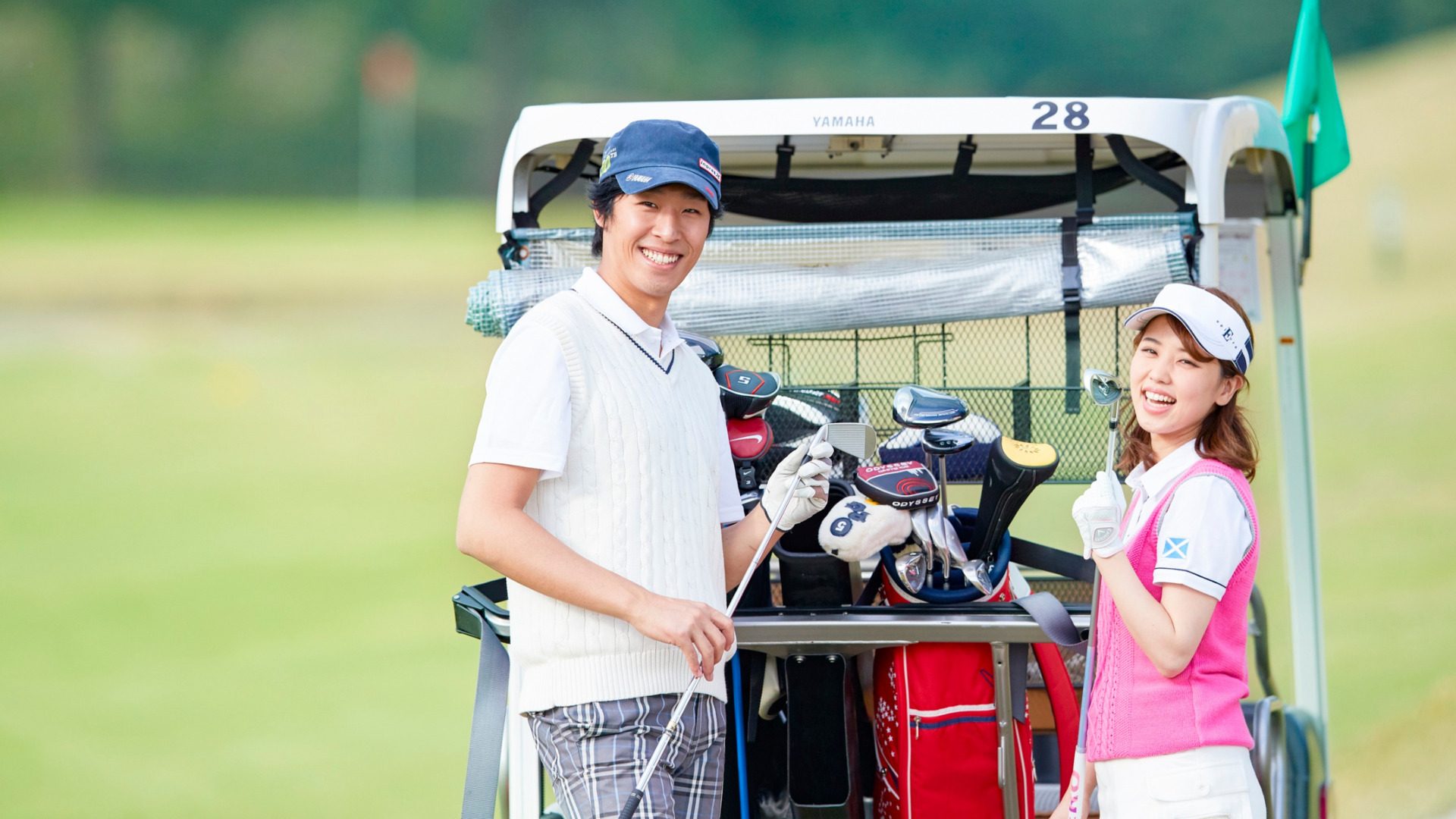 Two golfers smiling beside a golf cart loaded with clubs on a golf course.